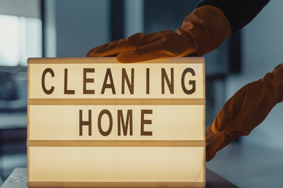 Close-up image of a household cleaning brush with beige plastic bristles and a wooden handle, placed inside a black dustpan on a wooden floor. The floor surface appears clean and well-maintained, with